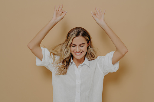 Carefree lovely female in white shirt with blonde hair enjoying nice day, keeping eyes closed and raising arms, making okay gesture with both hands and demonstrating positive emotions