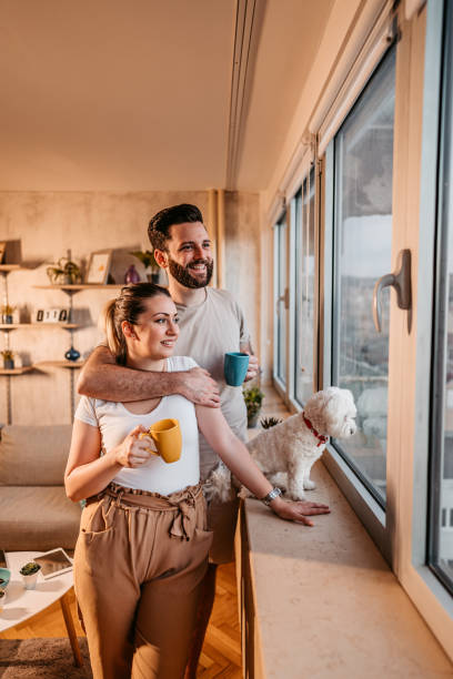 Young couple and dog looking through the window in their home Young lovely heterosexual couple and toy dog standing by the window in their home and looking through window. two dogs hugging stock pictures, royalty-free photos & images