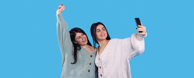 Cheerful twin girls making selfie isolated on blue background one of them raised her right hand up and smiling. High quality photo