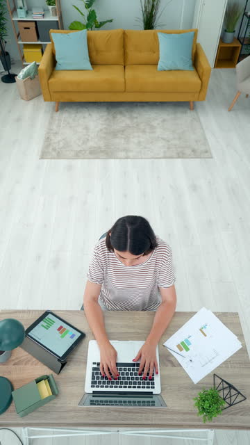 Young woman sitting at her desk and working remotely. She is planning new ideas for a new project or doing home finances using a laptop and financial reports. Vertical scene.