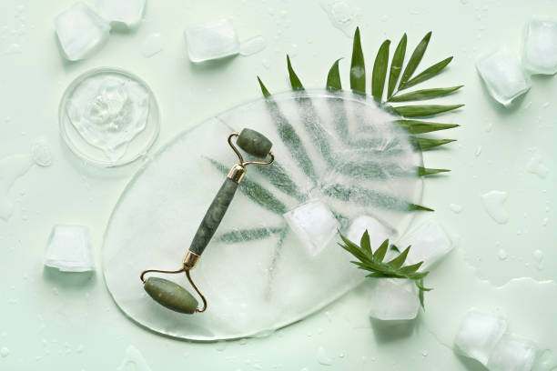 istock Self made moisturizer and green jade face roller on oval piece of ice with ice cubes. Exotic palm leaves on mint green background. Minimal flat lay, top view. Facial massage, handmade cosmetics 1329552010