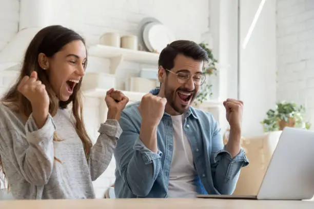 Overjoyed mixed race couple sit in kitchen looking at laptop screen celebrate online lottery victory. Happy young spouses read great news feeling amazed scream with joy enjoy big sale discount concept