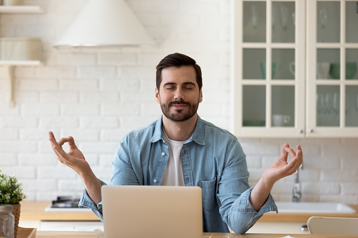 35s handsome man take break from telecommute on laptop, make meditation practice sit in kitchen relaxing with eyes closed. No stress, good life habits, fatigue tiredness relief, yoga lifestyle concept