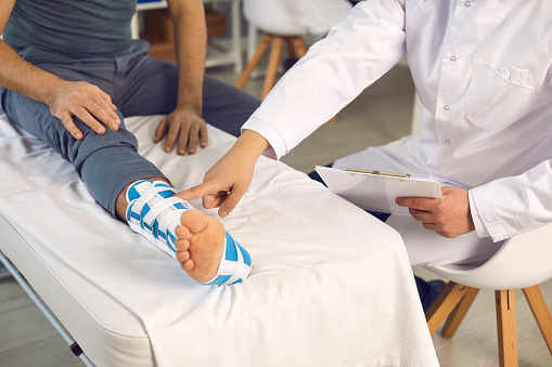Male doctor examine patient with injured broken leg on hospital bed. Midsection specialist point to bandage. Traumatologist, chiropractic appointment in clinic office for trauma treatment and recovery