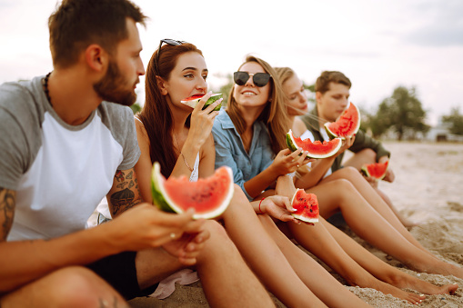 Young friends relaxing on the beach and eating watermelon. Group of young sitting together sitting near the sea and  enjoy summer party. People, lifestyle, travel, nature and vacations concept.