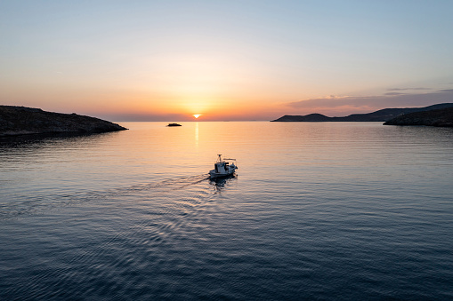 Fishing boat at sunrise. Cyclades island. Greece. Aerial drone view of an outbound typical wooden fishing boat moving on calm sea and orange color sky background.