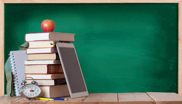 istock Stack of books with red apple, school bag, white notebook, silver clock, coloured pencils and digital tablet on the wood table in the chalkboard background 1329566478