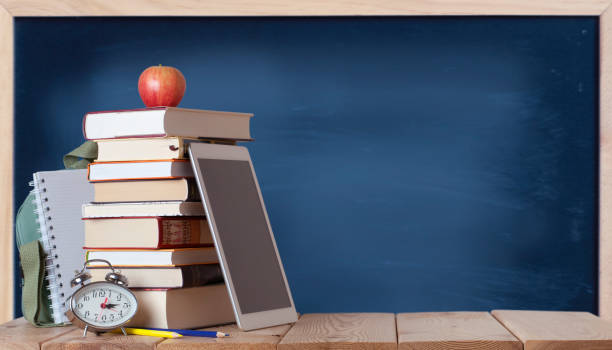 istock Stack of books with red apple, school bag, white notebook, silver clock, coloured pencils and digital tablet on the wood table in the chalkboard background 1329566479