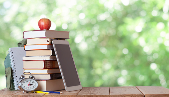 Stack of books with red apple, school bag, white notebook, silver clock, coloured pencils and digital tablet on the wood table in the nature blurred background with bokeh lights and copy space