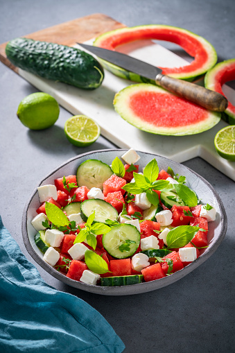 Healthy vegetarian watermelon salad bowl with feta cheese, cucumber and parsley on gray background with cutting board and knife, Mediterranean diet