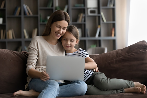 Happy gen Z daughter kid and smiling millennial young mom using laptop on sofa together, shopping on internet, watching movie, online video, enjoying leisure, relaxing with digital device at home
