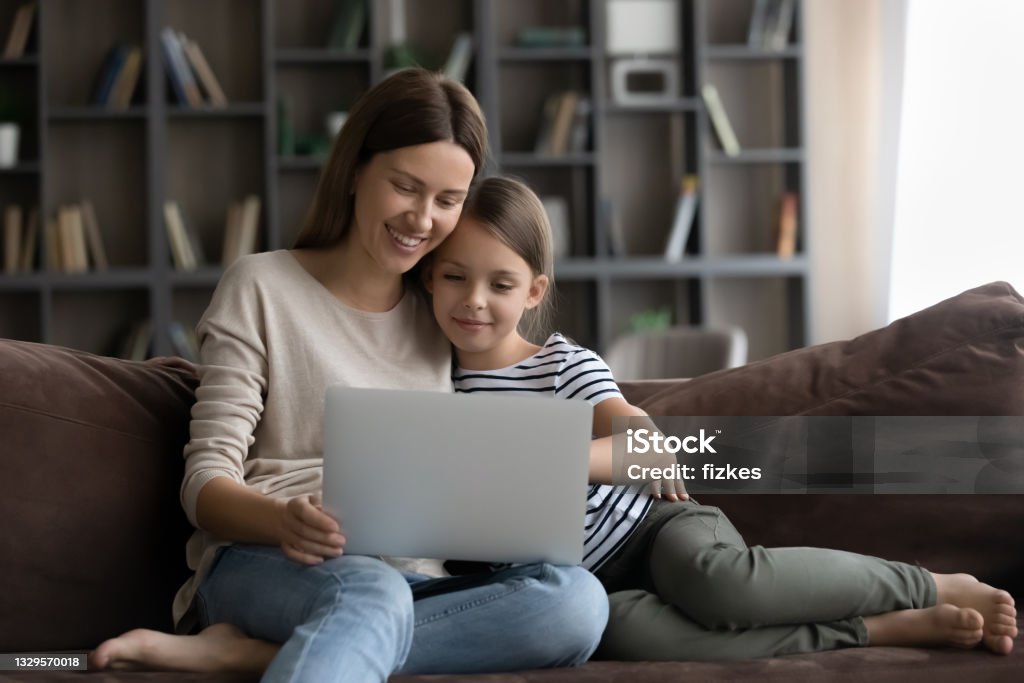 Happy daughter kid and smiling young mom using laptop Happy gen Z daughter kid and smiling millennial young mom using laptop on sofa together, shopping on internet, watching movie, online video, enjoying leisure, relaxing with digital device at home Mother Stock Photo