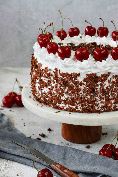istock Close-up image Black Forest gateau on cake stand, piped whipped cream rosettes with morello cherries, luxury chocolate cake frosted with whipped cream and covered with chocolate shavings, grey muslin, knife, marble effect background, focus on foreground 1329586668