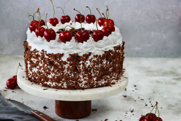 istock Close-up image Black Forest gateau on cake stand, piped whipped cream rosettes with morello cherries, luxury chocolate cake frosted with whipped cream and covered with chocolate shavings, grey muslin, knife, marble effect background, focus on foreground 1329586699