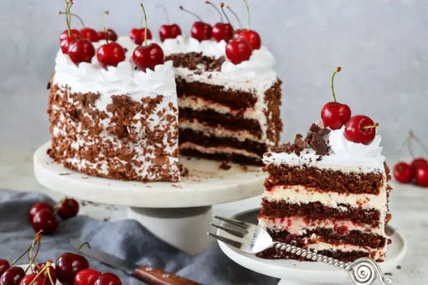 Photo of Image of sliced Black Forest gateau on cake stand with grey muslin, piped whipped cream rosettes topped with morello cherries and covered with chocolate shavings, beside slice of cake on a plate, marble effect background