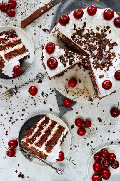 istock Image of Black Forest gateau on cake stand, piped whipped cream rosettes topped with morello cherries, luxury chocolate cake frosted with whipped cream and covered with chocolate shavings, grey muslin, knife, marble effect background, elevated view 1329587534