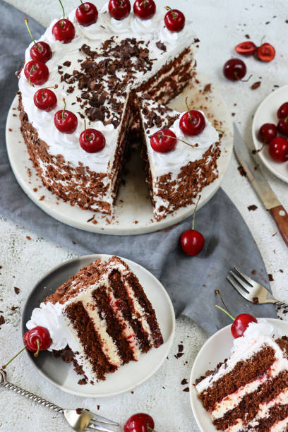 istock Image of Black Forest gateau on cake stand, piped whipped cream rosettes topped with morello cherries, luxury chocolate cake frosted with whipped cream and covered with chocolate shavings, grey muslin, knife, marble effect background, elevated view 1329587543