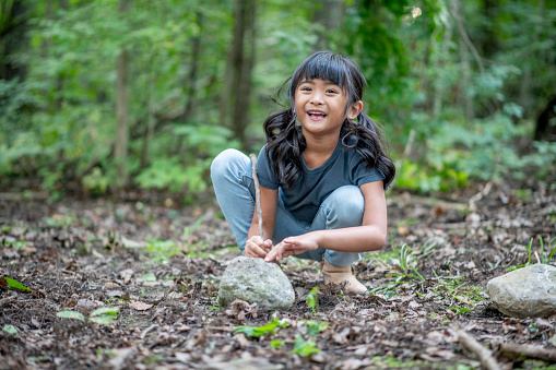 A girl crouches on the forest floor, smiling while she learns to make a fire.