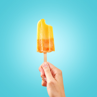 Woman holds bitten ice cream on blue background with copy space. Color frozen fruit popsicle.