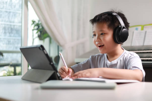 Boy using tablet computer doing homework Portrait of little Asian boy using tablet computer and wireless headphones at home, e-learning and home education concept. asian kids learning stock pictures, royalty-free photos & images