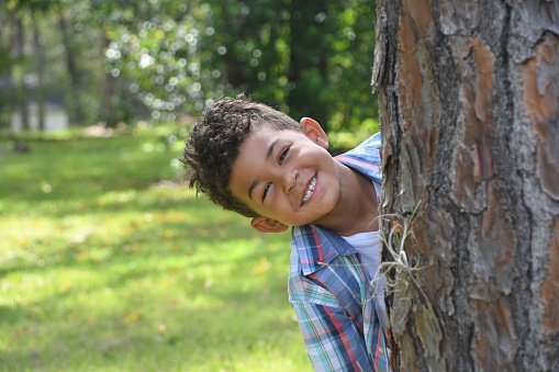 A biracial boy peeks out from behind a tree with a smile on his face while playing outside.