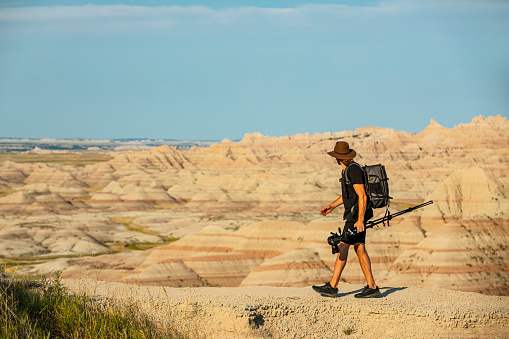 Photographer hiking through desert badlands wearing black backpack and holding tripod on bright sunny day