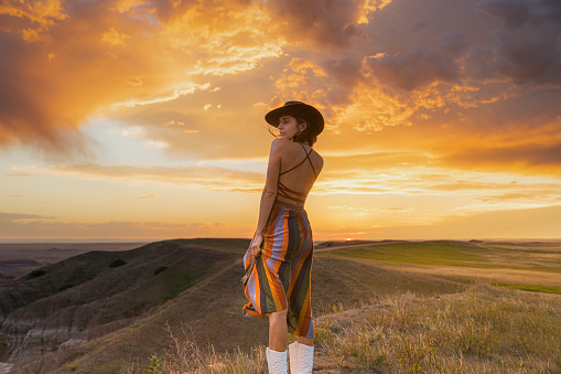 Beautiful woman in wide open field wearing multicolor dress standing in front of golden sunset