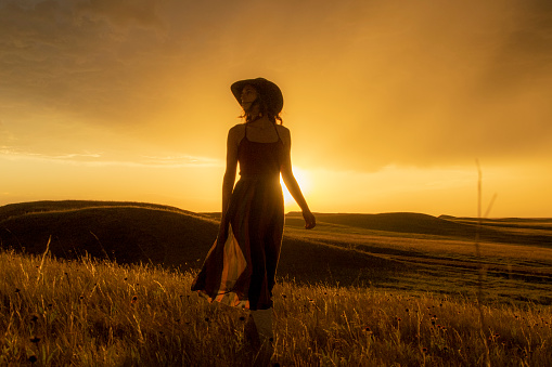 Woman in cowboy hat and dress walking along golden meadow at sunset