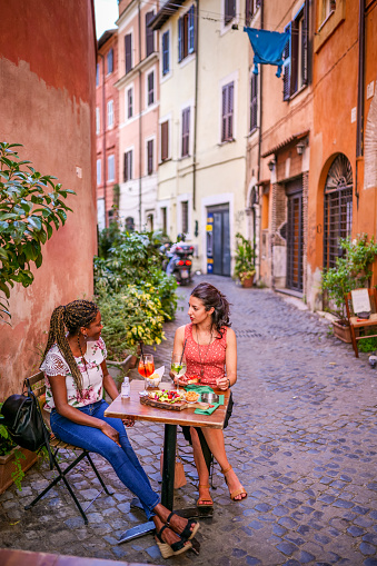 Rome, Italy, July 15 -- A couple of young and mixed race women enjoy Italian cuisine sitting outside a lovely and characteristic restaurant and winery in the ancient Trastevere district in the heart of Rome, near Sant'Egidio square. Trastevere is an iconic neighborhood of the Eternal City, due to the presence of countless artistic and historical treasures, monuments and ancient Romanesque and Baroque churches, but also for its squares and alleys to explore, where it is easy to find typical restaurants, pubs, small shops craftsmanship and the original Roman soul. In 1980 the historic center of Rome was declared a World Heritage Site by Unesco. Image in high definition format.