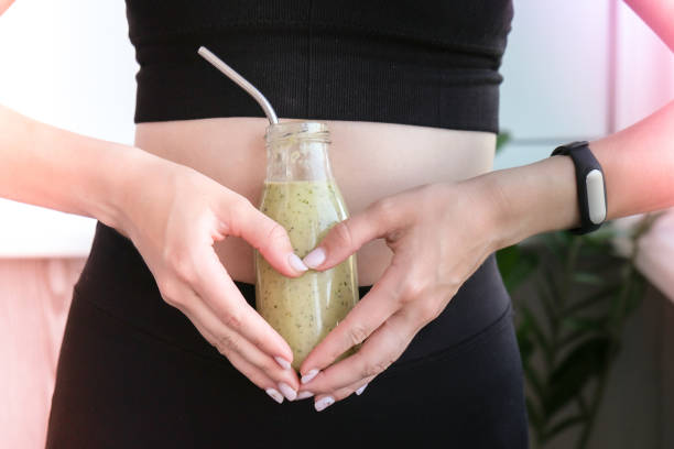 Hand in heart symbol holding smoothie drink detox on fit and slim belly woman. Fitness wellbeing concept. Body care, clean eating, weight loss diet concept. Woman with bowel or reproductive problem Hand in heart symbol holding smoothie drink detox on fit and slim belly woman. Fitness wellbeing concept. Body care, clean eating, weight loss diet concept. Woman with bowel or reproductive problem. Working out at home. Belly waist exercises detox stock pictures, royalty-free photos & images