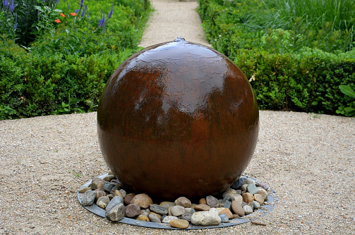veronica spicata, tilia cordata, buxus sempervirens, ornamental boxwood hedge and beige paths in the park. a stone ceramic ball from which water flows, like a calm fountain. symmetrical composition with blue perennials in the garden