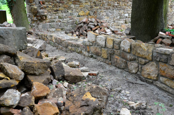 an old stone wall is being reconstructed between the two plots. the stones are dismantled and re-glued to the cement mortar. stonemason builds a wall in the park. the joints are repaired and the bushes are covered with a protective fabric, hedera helix se está reconstruyendo un antiguo muro de piedra entre las dos parcelas. las piedras se desmontan y se vuelven a pegar al mortero de cemento. cantero construye una pared en el parque. las juntas se reparan y los arbustos se cubren con una tela protectora