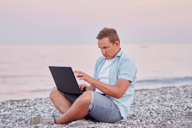 istock Man brunette with a laptop on a on the beach. Freelancer, working remotely, webinar, learning concept, chatting with friends. New normal concept 1329635739