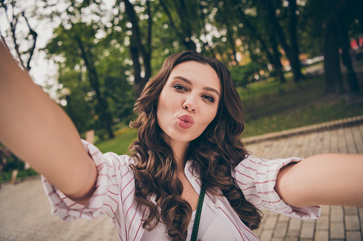 Photo portrait of pretty young girl with long wavy hair taking selfie sending air kiss with pouted plump lips in city park.
