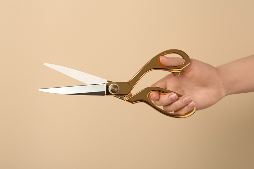 Woman holding sewing scissors on beige background, closeup