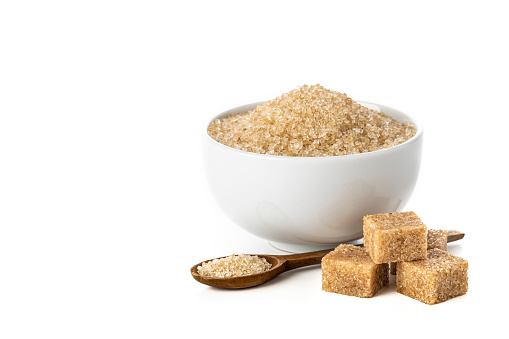 Front view of a bowl full of brown sugar beside some sugar cubes and a teaspoon. Objects are isolated on white background.