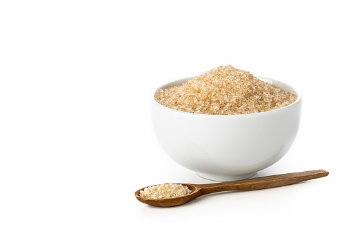 Front view of a bowl full of brown sugar beside a teaspoon. Objects are isolated on white background.