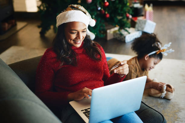 aufnahme einer jungen frau mit laptop und kreditkarte, während ihre tochter zu weihnachten zu hause im hintergrund spielt - christmas shopping internet family stock-fotos und bilder