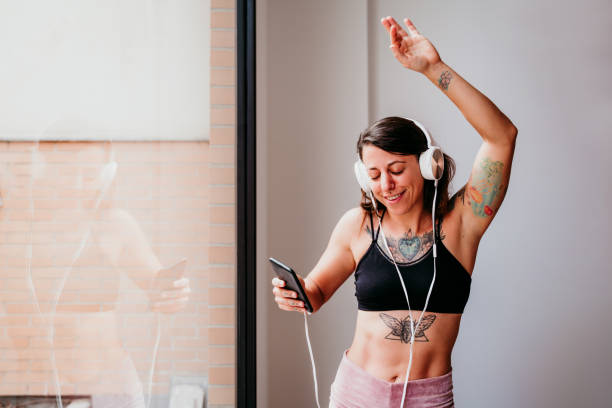 junge muskulöse kaukasische frau, die musik auf handy und headset im fitnessstudio hört. tagsüber am fenster tanzen. sport und gesunder lebensstil - headphones music mobile phone tattoo stock-fotos und bilder