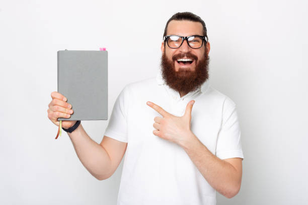 istock Man in glasses with beard holds grey book. Guy with closed notebook points at it over white background 1329652623