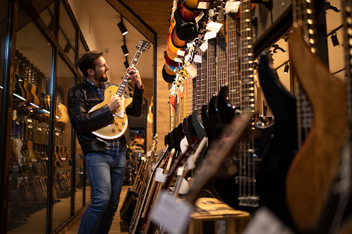 Proud young rocker musician in leather jacked playing electric guitar in music shop.