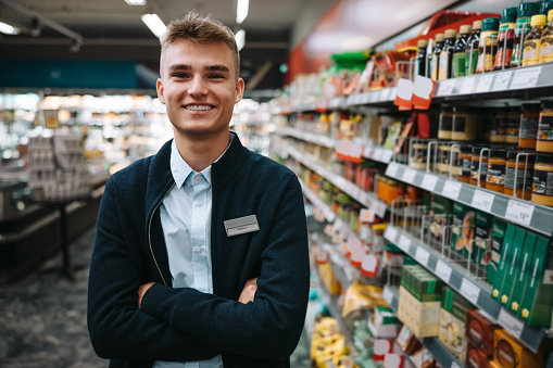 Portrait of a man in uniform standing at with arms crossed at grocery store. Male worker at supermarket.