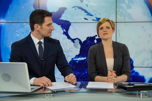 Man and woman sitting at news desk in a tv recording studio. Preparing to go live.