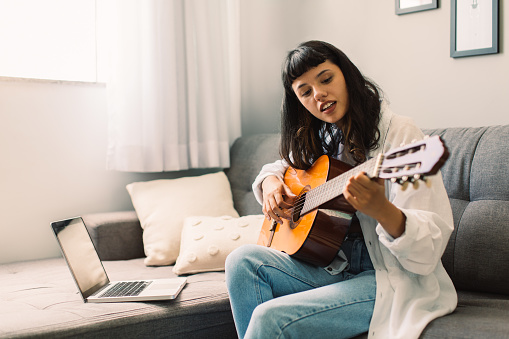 Female musician teaching a guitar lesson on her laptop. Young female guitarist giving an online music lesson on a video call. Woman teaching music at home during quarantine.