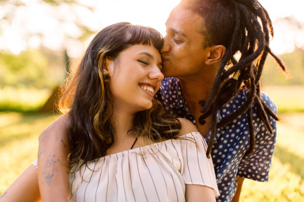 Beautiful couple being romantic outdoors Beautiful young interracial couple being romantic while sitting in a park. Affectionate young man kissing his girlfriend on her forehead while embracing her during the day. couple relationship stock pictures, royalty-free photos & images