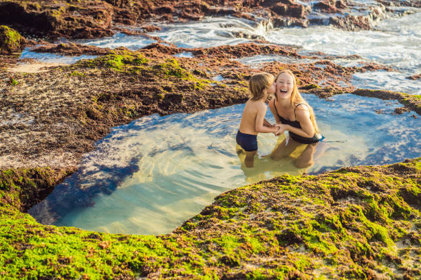 turistas de mãe e filho na praia pantai tegal wangi sentados em um banho de água do mar, ilha bali, indonésia. bali travel concept. viajando com o conceito de crianças. lugares amigáveis para crianças - kuta beach - fotografias e filmes do acervo