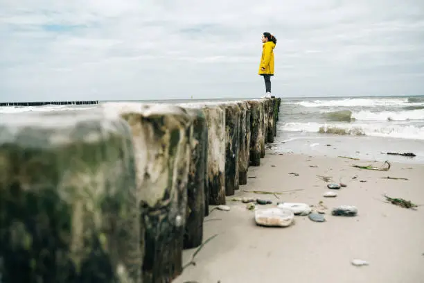 little girl in yellow raincoat standing on groyne at beach and watching the baltic sea