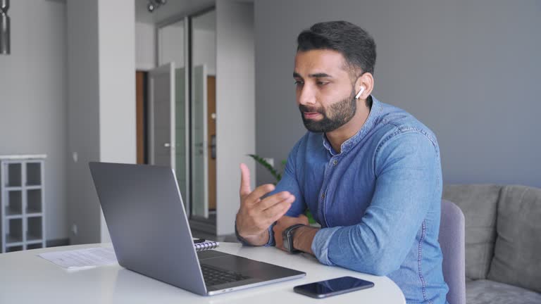 Adult indian man wearing wireless earphones make video call looking at laptop