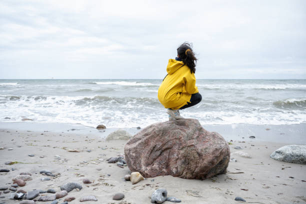 little girl in yellow raincoat crouching on stone watching the sea rear view on little girl in yellow raincoat sitting on stone watching the baltic sea baltic sea people stock pictures, royalty-free photos & images