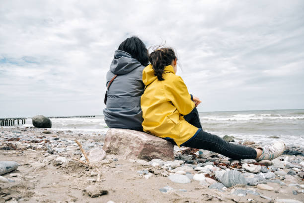 little girl in yellow raincoat sitting with mother on stone at beach rear view on little girl in yellow raincoat sitting with mother at beach on stone and watching the baltic sea baltic sea people stock pictures, royalty-free photos & images
